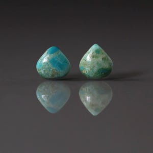May include: Two teardrop-shaped turquoise gemstones with a mottled pattern of blue and green. The stones are set against a dark gray background, with their reflections visible below. The gemstones are likely for jewelry making.