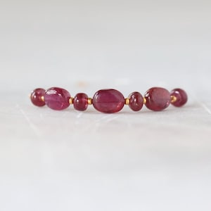 May include: A bracelet featuring a series of polished, oval-shaped deep pink gemstones arranged in a row. The stones are separated by small, round gold-colored metal beads, creating a consistent pattern along the length of the jewelry piece.