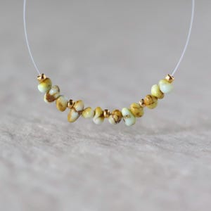 May include: A necklace featuring a series of small, irregular, pale green and brown mottled stone beads interspersed with tiny, round, metallic gold-colored spacer beads, all strung on a thin, clear, flexible monofilament cord.