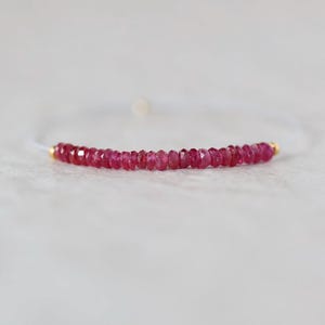 May include: A delicate bracelet featuring a single row of small, faceted, deep pink ruby beads strung on a thin, translucent cord. The strand of beads is accented by two tiny, polished gold-toned metal beads at each end of the gemstone section.