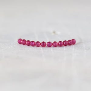May include: A delicate bracelet featuring a single row of small, round, faceted beads in a vibrant magenta-pink color. The beads are strung together in a horizontal line, showcasing a uniform size and a reflective, sparkling surface texture.
