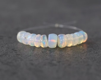 Könnte beinhalten: Ein Armband aus durchscheinenden, abgerundeten Opalperlen, die auf einen dünnen, klaren Faden aufgezogen sind. Die Perlen haben ein milchig weißes Aussehen mit subtilen irisierenden Blitzen in Blau, Orange und Gelb auf ihren glatten Oberflächen.