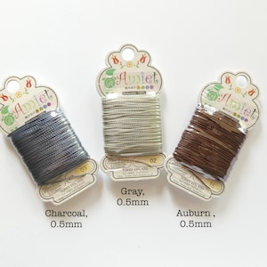May include: Three spools of Amiet brand thread in charcoal, gray, and auburn. Each spool is labeled with the color name and 0.5mm thickness. The packaging includes the text "Polyester 100%" and Japanese text.