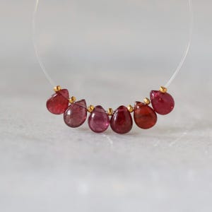 May include: A necklace featuring six teardrop-shaped pink and red gemstones strung on a clear filament. The stones are separated by small, round, metallic gold-toned beads, creating a delicate, curved arrangement of polished, translucent gems.