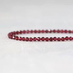 May include: A single strand of small, deep red garnet beads arranged in a gentle curve. Each bead features a faceted surface that reflects light, creating a consistent, sparkling appearance across the entire length of the delicate jewelry piece.