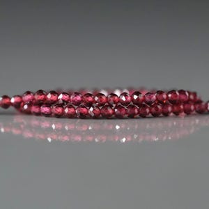 May include: Strands of faceted, deep red garnet beads. The beads are small and round, with multiple facets that catch the light. The strands are arranged in a layered pattern, creating a sense of depth and texture. The background is a soft, neutral gray.