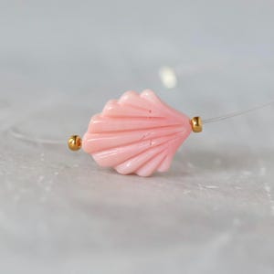 May include: A necklace featuring a single light pink seashell-shaped bead centered on a clear nylon cord. The seashell bead is flanked by two small, round, metallic gold-colored spacer beads, creating a simple and delicate jewelry piece.