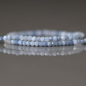 May include: Two strands of light blue faceted gemstone beads. The beads are small and round, with a slightly iridescent quality. The strands are arranged in a gentle curve, with a blurred background.