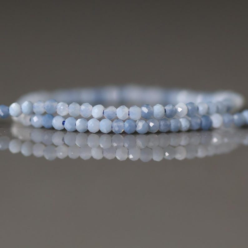 May include: Two strands of light blue faceted gemstone beads. The beads are small and round, with a slightly iridescent quality. The strands are arranged in a gentle curve, with a blurred background.