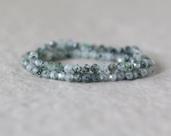 Könnte beinhalten: Ein Perlenarmband aus kleinen, facettierten, durchscheinenden hellgrünen Steinen mit dunkelgrünen und schwarzen Einschlüssen. Die Perlen sind in einer kreisförmigen Schleife angeordnet und zeigen eine reflektierende, facettierte Oberflächenstruktur.