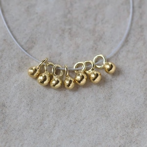 May include: A collection of seven small, gold-toned spherical charms, each featuring a circular loop at the top for attachment. The charms have a textured, slightly irregular surface finish and are strung together on a thin, clear filament wire.