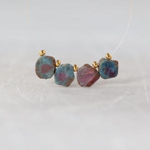May include: A necklace strand featuring four raw, irregularly shaped stones with blue and magenta hues, interspersed with small, polished gold-toned metal beads on a thin, clear filament string. The stones display natural textures and variations.