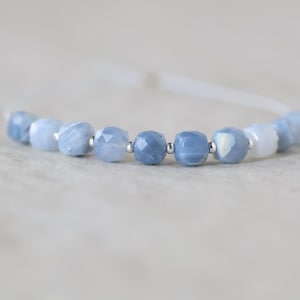 May include: A bracelet featuring a series of faceted, light blue, semi-translucent beads arranged in a row. Small, round, metallic silver-colored spacer beads are positioned between each of the blue beads along the thin, light-colored cord.