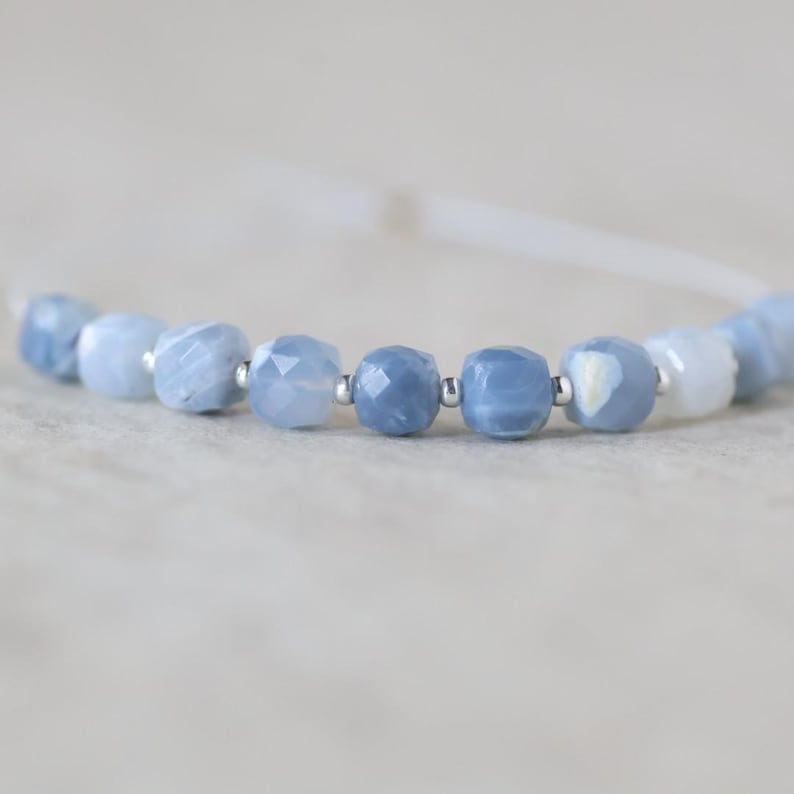May include: A bracelet featuring a series of faceted, light blue, semi-translucent beads arranged in a row. Small, round, metallic silver-colored spacer beads are positioned between each of the blue beads along the thin, light-colored cord.