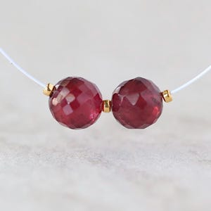 May include: Two faceted, deep red, round beads strung on a thin white cord. The beads are separated and flanked by small, polished gold-toned metal spacers. The design is simple, featuring two prominent dark red stones with a reflective surface.
