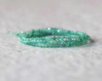 Puede incluir: Una pulsera de múltiples hilos compuesta por pequeñas cuentas verdes translúcidas y facetadas. Las cuentas están ensartadas en una disposición circular, creando una apariencia en capas con un color verde claro uniforme y una superficie reflectante.
