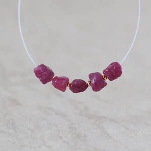 May include: A string of five raw, deep pink ruby gemstones arranged on a thin white cord. The rough-cut rubies are separated by small, polished gold-toned metal spacer beads, creating a linear sequence of alternating stones and metallic accents.