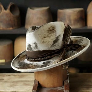 Può includere: Un cappello da cowboy bianco e marrone invecchiato con una fascia marrone e dettagli di piume. Il cappello è esposto su un ceppo di legno. La tesa è larga e la corona è piegata. Il cappello ha un'estetica vintage.