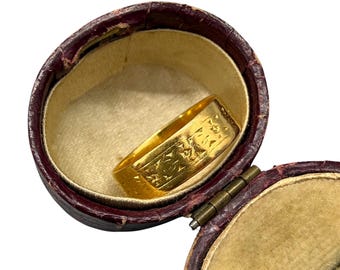 May include: A gold ring with an engraved design rests inside a vintage, burgundy leather jewelry box with a gold interior. The ring is rectangular and features intricate patterns.