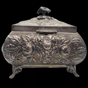 May include: A silver-plated trinket box with a hinged lid and ornate floral design. The box has a raised floral design with three roses and leaves. The box has four feet and a small knob on the lid.