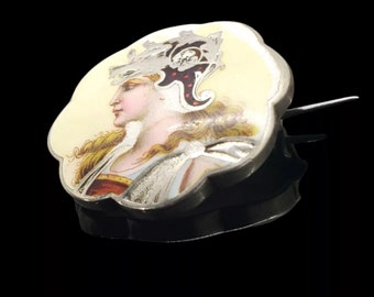Könnte beinhalten: Eine Vintage-Haarspange aus silberfarbenem Metall mit einem bemalten Emaille-Design. Das Emaille zeigt das Profil einer Frau mit blondem Haar, die einen Helm trägt. Die Spange hat einen schwarzen Sockel und einen dekorativen, gewellten Rand.