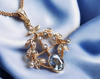 Könnte beinhalten: Eine goldfarbene Anhängerkette mit Blattdesign, verziert mit kleinen Perlen und einem großen, ovalförmigen, hellblauen Edelstein. Die Kette liegt auf blauem Satin.