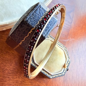 Pode incluir: Uma pulseira de ouro com uma fileira de pequenas pedras preciosas vermelhas. A pulseira é exibida em uma caixa de joias azul escura antiga com acabamento dourado. A caixa está aberta, revelando um interior amarelo claro.