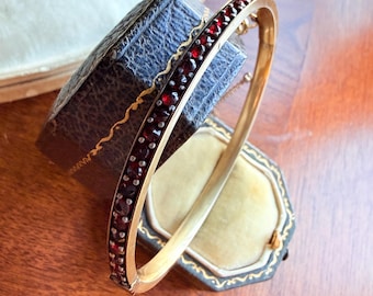 Puede incluir: Un brazalete de oro con una fila de pequeñas piedras preciosas rojas. El brazalete se exhibe en un joyero antiguo azul oscuro con ribete dorado. La caja está abierta, revelando un interior amarillo claro.
