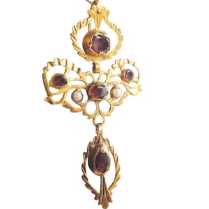 Puede incluir: Un collar con colgante dorado con un diseño de filigrana detallado. El collar presenta varias piedras preciosas de color rojo intenso y pequeñas perlas blancas. El colgante tiene una estética ornamentada y vintage, con un elemento en forma de lágrima en la parte inferior.