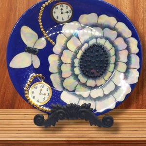 May include: A blue ceramic plate with a white flower design and two gold pocket watches with chains. The plate is on a black stand.