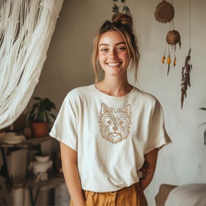 A smiling young freckled woman standing indoors in a cozy boho room, wearing an off-white t-shirt and brown pants. The chest of the t-shirt features a detailed golden-brown line art embroidery design of a Yorkshire Terrier dog's face. Digital machine embroidery file, apparel mockup, casual pet lover fashion.