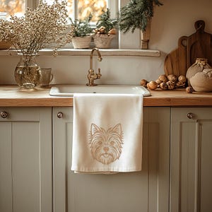A beige kitchen towel hanging from a white kitchen sink counter in a cozy rustic kitchen. The towel features a detailed golden-brown line art embroidery design of a Yorkshire Terrier dog's face. Digital machine embroidery file, home decor mockup, kitchen textile pet lover style.