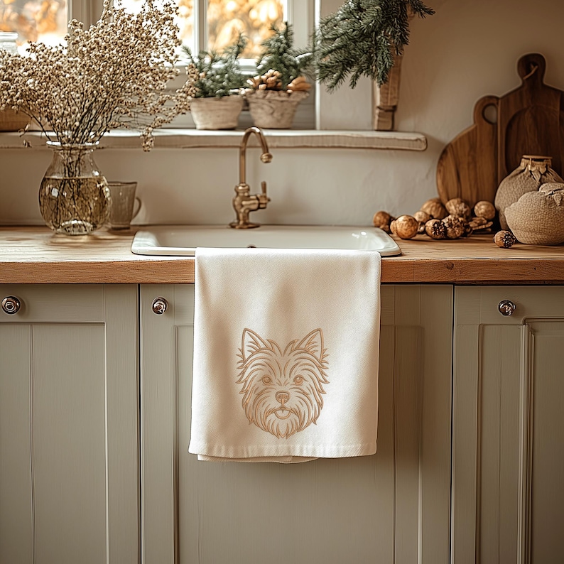 A beige kitchen towel hanging from a white kitchen sink counter in a cozy rustic kitchen. The towel features a detailed golden-brown line art embroidery design of a Yorkshire Terrier dog's face. Digital machine embroidery file, home decor mockup, kitchen textile pet lover style.