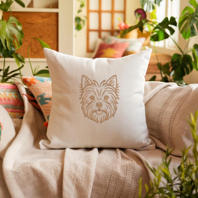 A white throw pillow resting on a cozy sofa surrounded by plants and blankets. The front of the pillow features a detailed golden-brown line art embroidery design of a Yorkshire Terrier dog's face. Digital machine embroidery file, home decor mockup, pet lover style.