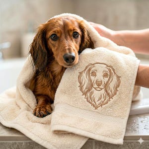 May include: A long-haired dachshund wrapped in a beige cotton towel. The towel features an embroidered line art illustration of a long-haired dachshund face on the front, matching the appearance of the dog wrapped within the fabric.