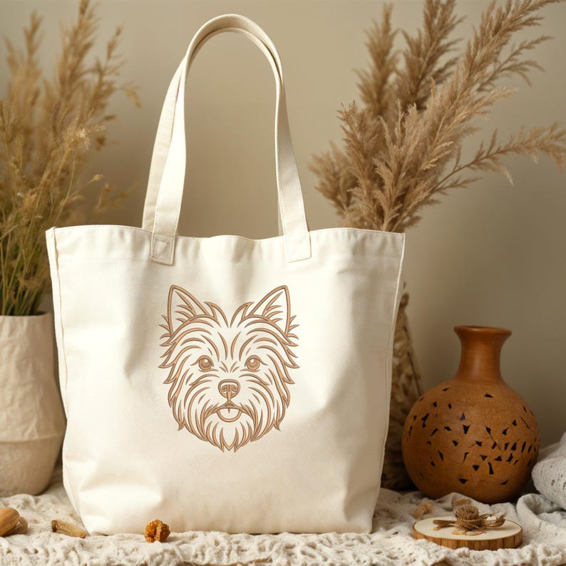 A beige canvas tote bag displayed on a neutral surface with dried plants and a decorative clay vase in the background. The front of the bag features a detailed golden-brown line art embroidery design of a Yorkshire Terrier dog's face. Digital machine embroidery file, accessory mockup, pet lover style.