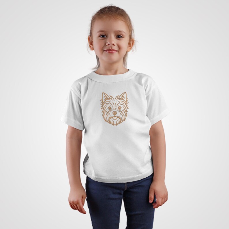 A cute young girl standing against a plain light gray background, wearing a white t-shirt and blue jeans. The chest of the t-shirt features a minimalist line art embroidery design of a Yorkshire Terrier dog's face in golden brown thread. Digital machine embroidery file, kids apparel mockup, pet lover style.