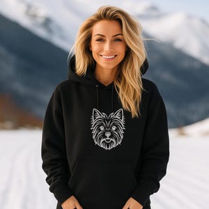 A smiling young blonde woman standing outdoors in a snowy mountain landscape, wearing a black hoodie. The front of the hoodie features a minimalist white line art embroidery design of a Yorkshire Terrier dog's face. Digital machine embroidery file, apparel mockup, winter casual pet lover fashion.