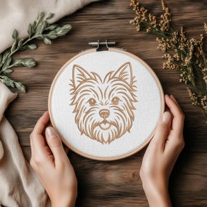 A person holding a round wooden embroidery hoop on a dark wooden background decorated with fabric and dried plants. Inside the white fabric, there is a golden-brown line art embroidery design of a Yorkshire Terrier dog's face. Digital machine embroidery file, craft mockup.