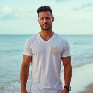 A handsome man standing on a beach near the ocean, wearing a white v-neck t-shirt. The chest of the t-shirt features a detailed golden-brown line art embroidery design of a Yorkshire Terrier dog's face. Digital machine embroidery file, apparel mockup, men's summer pet lover fashion.