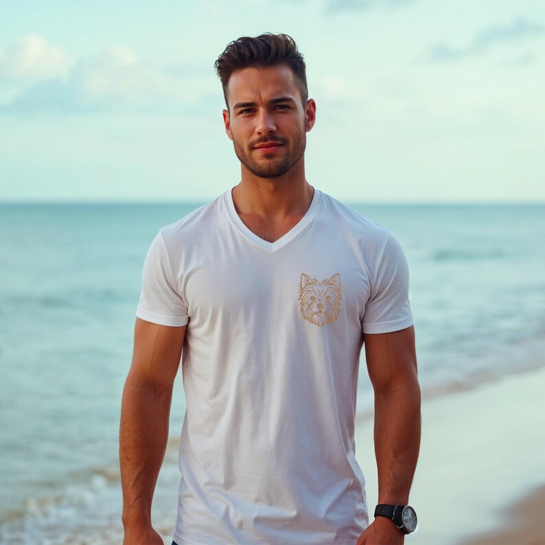 A handsome man standing on a beach near the ocean, wearing a white v-neck t-shirt. The chest of the t-shirt features a detailed golden-brown line art embroidery design of a Yorkshire Terrier dog's face. Digital machine embroidery file, apparel mockup, men's summer pet lover fashion.