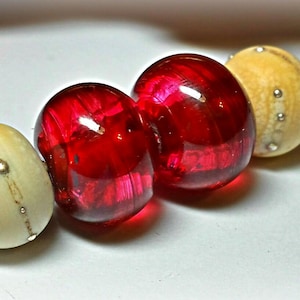 May include: A close-up of a decorative skewer featuring four beads. Two large, translucent red beads are flanked by two smaller, off-white beads with silver accents. The skewer has a natural, twig-like stem.