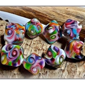 May include: A collection of colorful, square-shaped glass beads with swirling patterns in shades of red, blue, green, and yellow. The beads are displayed on a textured wooden surface, showcasing their unique, artistic designs. These beads are ideal for jewelry making.