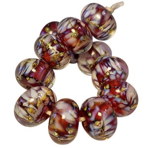 May include: A strand of artisan glass beads. The beads are a mix of red, white, and purple with gold flecks. The beads are strung together on a natural-colored cord. The beads are round and have a glossy finish.