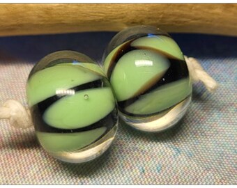 May include: Two glass beads with a light green core and black and brown swirling stripes. The beads are connected by a white string. The beads are oval-shaped and have a clear, glossy finish.