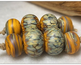 May include: A collection of handmade glass beads in shades of yellow, orange, blue, and purple. The beads have a swirled, marbled design and are strung on a natural fiber cord. These beads are perfect for jewelry making.