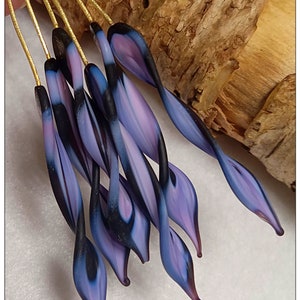 May include: Close-up of a collection of glass art pieces. Each piece features a slender, elongated shape with a gradient of colors, including shades of purple, blue, and black. The pieces are attached to thin, gold-colored wires, suggesting they are decorative items.