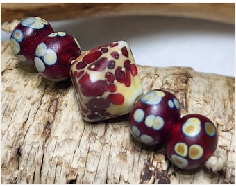 May include: A collection of handmade glass beads in shades of burgundy, cream, and yellow. The beads are round and square, with a speckled and spotted design. The beads are strung together on a natural fiber cord, perfect for jewelry making.
