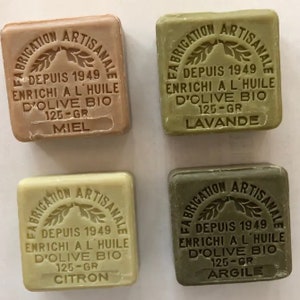 May include: Four bars of soap with different scents. Each bar is square and has a brown label with white text. The text on the labels reads "Fabrication Artisanale" and includes the scent of each bar: Miel, Lavande, Citron, and Argile. Each bar is 125 grams.