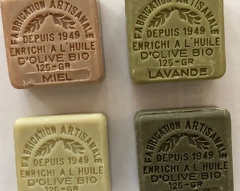 May include: Four bars of soap with different scents. Each bar is square and has a brown label with white text. The text on the labels reads "Fabrication Artisanale" and includes the scent of each bar: Miel, Lavande, Citron, and Argile. Each bar is 125 grams.